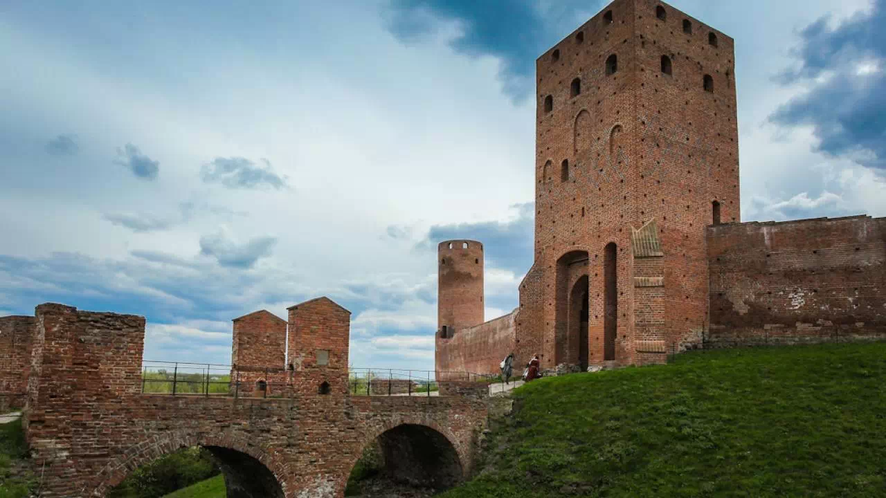 Zamek, a właściwie jego ruiny, o gotyckiej architekturze z wysoką, ceglano-murarską wieżą i mostem łukowym na zielonym terenie. Widok zamku otoczonego murami obronnymi i niebem pełnym chmur.