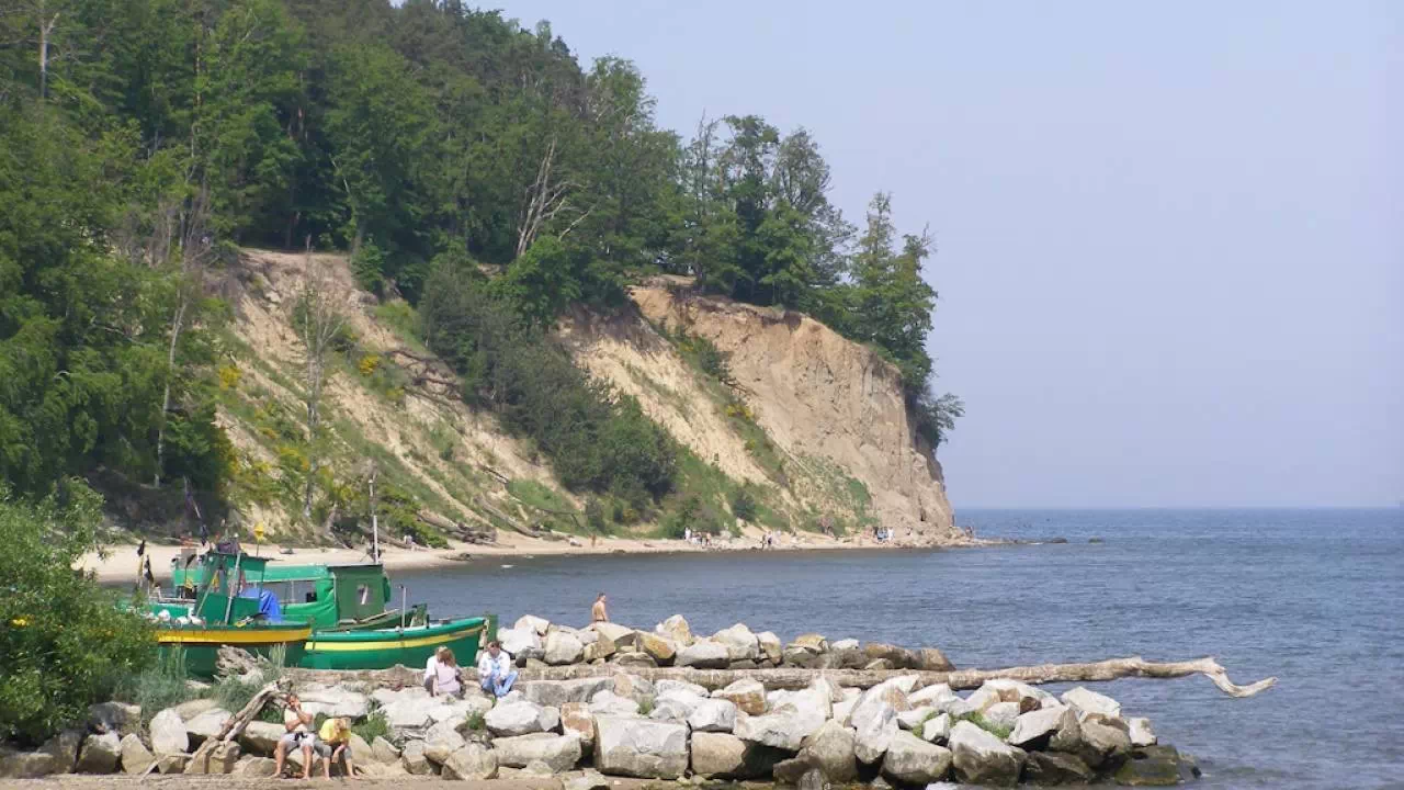 Zdjęcie przedstawiające zatokę Gdańską i Klif Orłowski. Na pierwszym planie falochron, zielona łódka rybacka oraz grupa turystów wypoczywających na słońcu