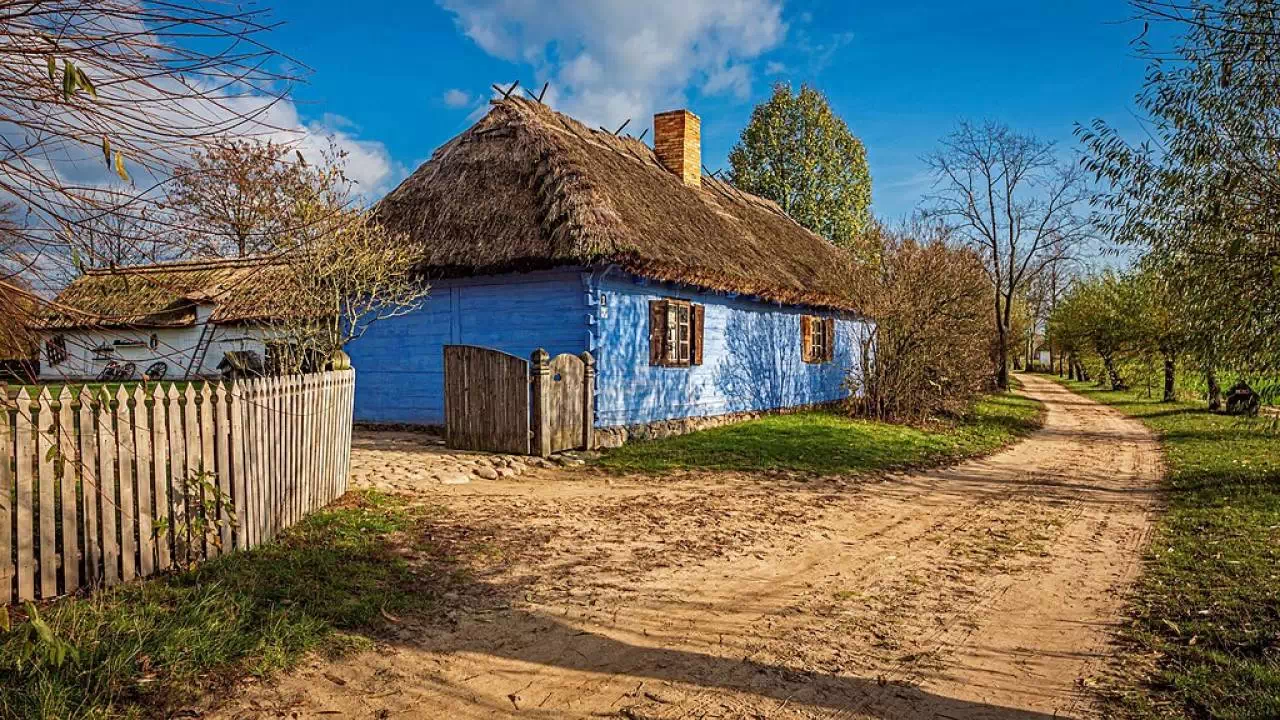 Tradycyjna, drewniana chata kryta strzechą, otoczona drewnianym płotem, z ogrodem pełnym zieleni.