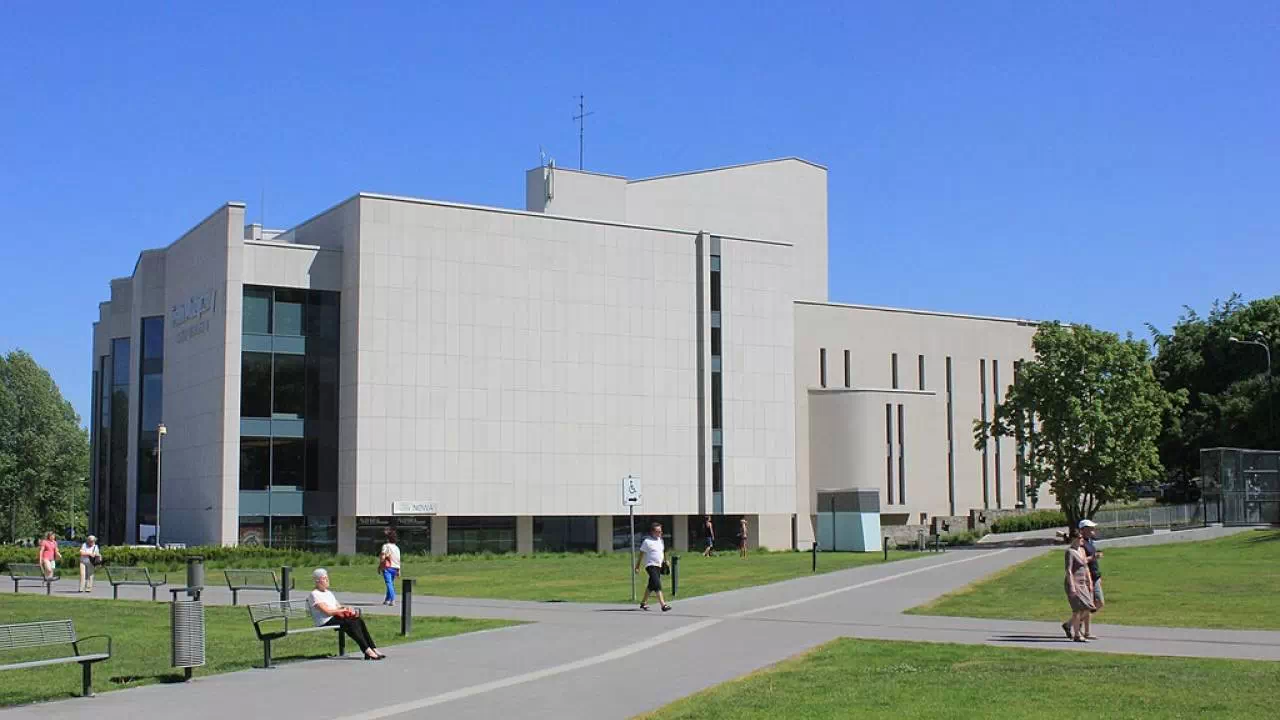 Budynek Teatru Muzycznego w Gdańsku. Szaro-beżowa konstrukcja otoczona skwerem, parkiem i zielenią.