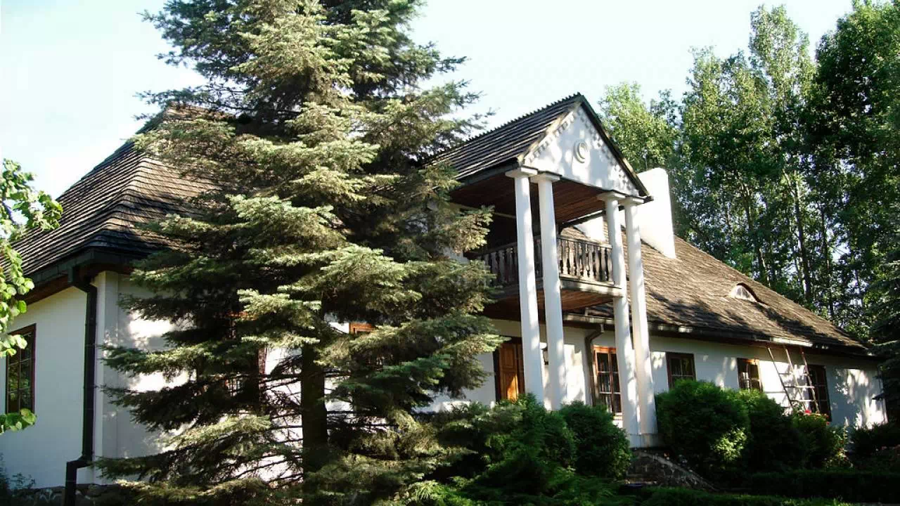 Drewniany wiejski dom kryty strzechą, otoczony zielenią, z kwitnącymi roślinami w przydomowym ogródku. Tradycyjna architektura wiejska, przedstawiająca zabudowę charakterystyczną dla dawnej polskiej wsi.