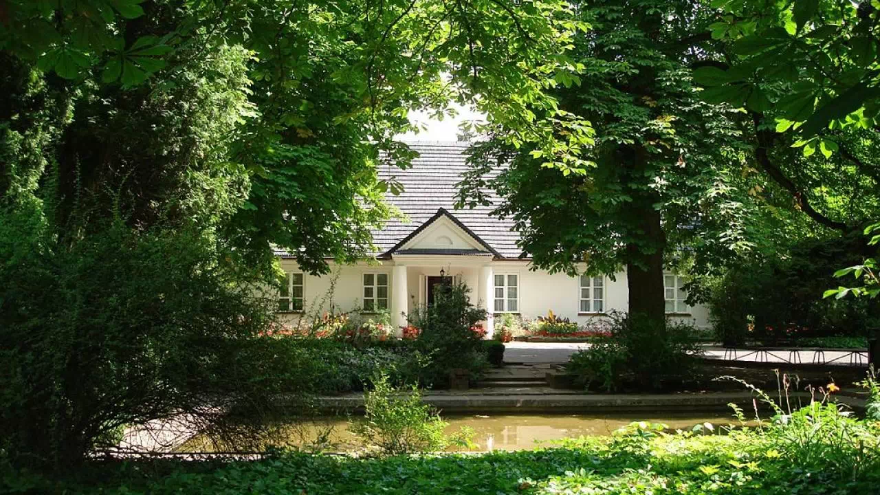 Budynek dworku w Żelazowej Woli. Minimalistyczny budynek z białą elewacją, kolumnadą przed wejściem i czarnym dachem. Przed nim znajduje się park pełen drzew i krzewów, a także staw.